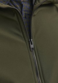 Povečava zadrge na temno zeleni jakni, delno odprta, razkriva modro-rjavo karo srajco spodaj.