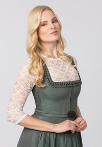 Die traditionelle Tracht weist ein grünes Mieder mit einem strukturierten Stoff auf, dazu gehören Spitzenärmel und geflochtene Verzierungen. Eine dekorative Brosche schmückt die Taille.