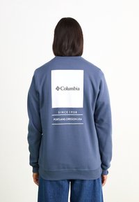 Granatowy sweter z miękkiego materiału, z białym logo w prostokątnym kształcie na plecach z napisem. Żebrowane mankiety i dół zapewniają dopasowane wykończenie.