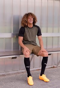 Conjunto atlético que incluye una camiseta de manga corta en verde oscuro y negro, pantalones cortos color beige, calcetas negras hasta la rodilla y botas de fútbol de color naranja brillante.
