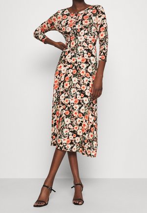 Femme portant une robe mi-longue noire à fleurs avec des manches trois-quarts et des talons noirs à bride à la cheville, debout avec une main sur la hanche.