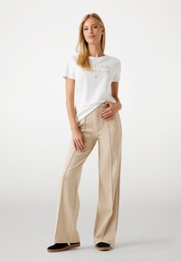 Pantalons larges beiges à taille haute, associés à un t-shirt blanc avec imprimé logo doré. Le modèle se tient avec une main sur la hanche.