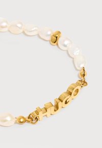 Βραχιόλι με ακανόνιστες λευκές πέρλες και χρυσά χάντρες με χρυσά γράμματα που σχηματίζουν τη λέξη «HUGO» σε λευκό φόντο.
