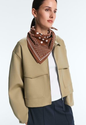 Femeie purtând o jachetă bej cu fermoar și croială scurtă, tricou alb, fustă bleumarin și o eșarfă maro cu buline legată în jurul gâtului, uitându-se spre dreapta.