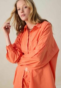 Chemise à manches longues corail à boutons avec une poche, offrant une coupe décontractée et une texture douce, assortie à un pantalon.