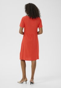 Robe rouge à manches courtes avec une coupe décontractée, présentant un design de dos plissé. Porteée avec des talons beige clair, sur un fond blanc uni.