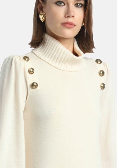 Maglione color crema con colletto alto a coste, maniche a sbuffo e bottoni dorati sulle spalle. Tessuto morbido e texture lavorata.