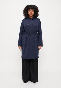 Marinblå trenchcoat med dubbelknäppning, bred krage, bälte i midjan och framfickor; matchad med svarta vida byxor.
