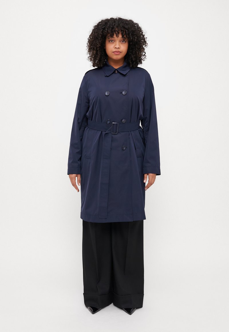 Marinblå trenchcoat med dubbelknäppning, bred krage, bälte i midjan och framfickor; matchad med svarta vida byxor.