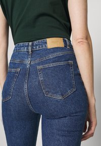 Jeans de mezclilla azul oscuro de talle alto con un parche de cuero marrón, diseño de cinco bolsillos y detalles de costura visibles en la parte trasera.