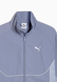 Svetlomodrá bunda Puma s vysokým golierom, celým zipsom, bielym logom na hrudi a jemnými textúrovanými detailmi pozdĺž švov.