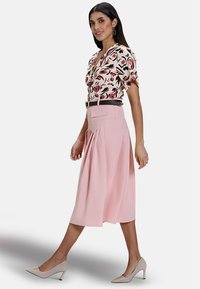 Blouse à motifs floraux avec des manches courtes, ceinture noire et jupe midi plissée rose. Des chaussures à talons clairs complètent la tenue.