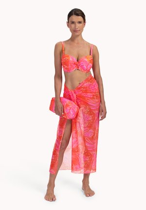Frau trägt ein Bikinioberteil mit auffälligem Pink- und Orange-Muster, dazu einen passenden Wickelrock mit Oberschnitt und hält eine ebenso gemusterte Clutch-Tasche.