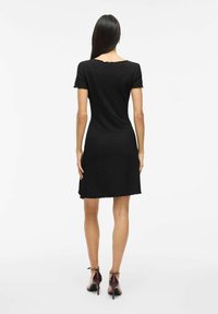 Vestido negro de manga corta con tejido texturizado, dobladillo con forma de ola y silueta ajustada. Llevado con zapatos de tacón alto marrones con tira en el tobillo.