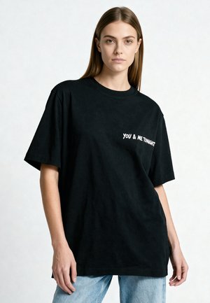 Jonge vrouw met steil bruin haar die een zwart oversized T-shirt met witte tekst "YOU & ME TONIGHT" en lichtblauwe jeans draagt.