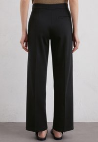 Schwarze weit geschnittene Hosen aus glattem Stoff mit einem nahtlosen Bund und einer hinteren Tasche, kombiniert mit flachen Sandalen.