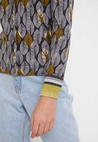 Strickjacke mit einem Blattmuster in Grau und Senf auf dunklem Hintergrund. Ärmelbündchen in Grün und Grau gestreift. Knopfschluss und strukturierter Stoff.