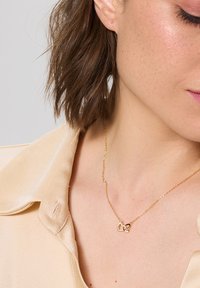 Naszyjnik w odcieniu złota z dwoma przeplatającymi się sercami, z których jedno zdobione jest małymi cyrkoniami, na tle jasnobeżowej jedwabnej koszuli.