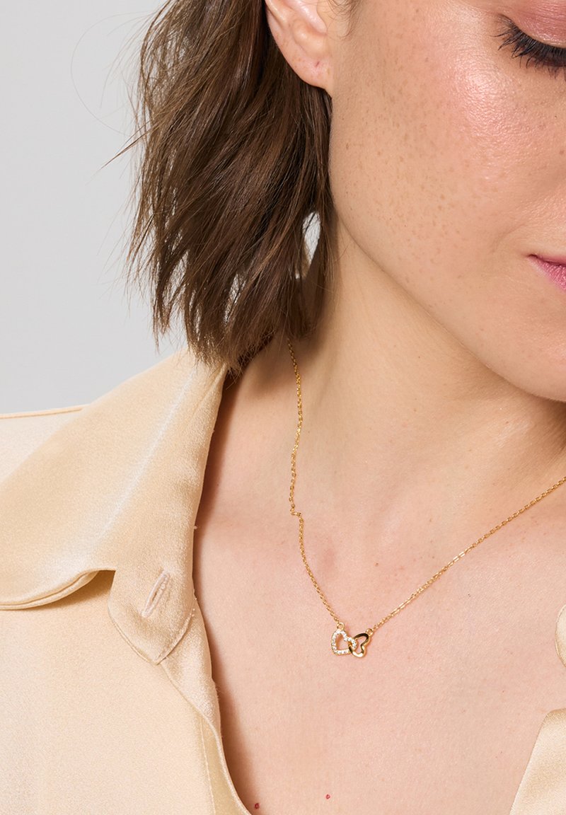 Naszyjnik w odcieniu złota z dwoma przeplatającymi się sercami, z których jedno zdobione jest małymi cyrkoniami, na tle jasnobeżowej jedwabnej koszuli.