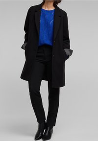 Schwarzer Wollmantel, strukturierter blau gestrickter Pullover, maßgeschneiderte schwarze Hose und spitze schwarze Stiefeletten. Graue Akzente an den Mantelärmeln.