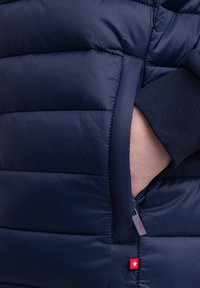 Gilet matelassé navy avec motif coussiné, doté d'une poche zippée sur le côté et de poignets côtelés, fabriqué en tissu lisse et léger. Détail du logo rouge.
