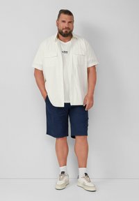Wit short-sleeve overhemd over een witte T-shirt, gecombineerd met marineblauwe shorts. Accessoires zijn witte sneakers met beige details.