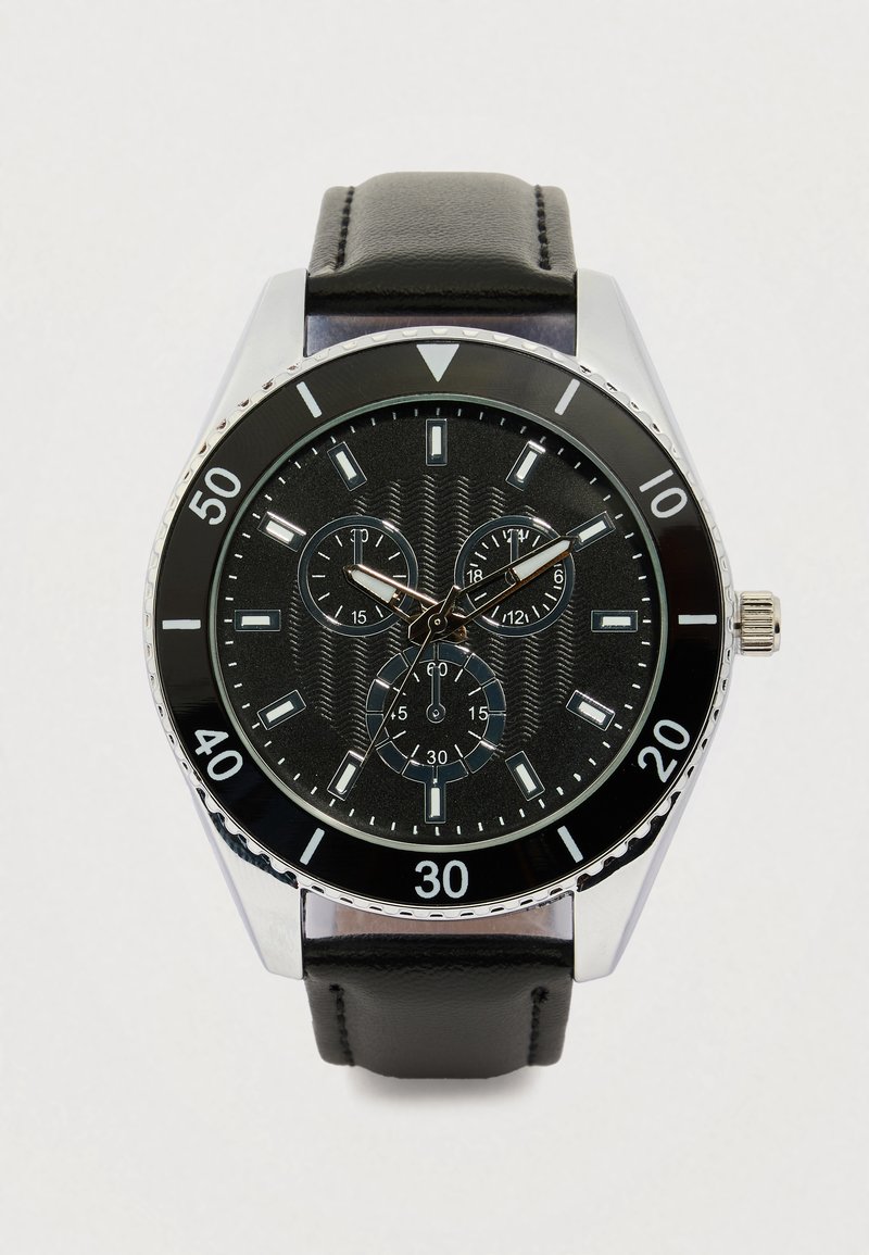 Ρολόι με μαύρο δερμάτινο λουρί, ασημένια και μαύρη στρογγυλή κάσα, υφασμένο μαύρο καντράν, τρεις υποκουρδίσματα και έντομους λευκούς δείκτες ώρας.