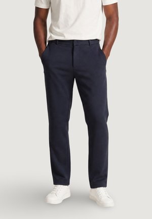 Homme portant un pantalon bleu marine, des baskets blanches et une chemise blanche à manches courtes, les mains dans les poches, debout sur un fond uni.