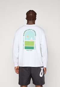 Maglietta a maniche lunghe bianca con una grafica colorata sul retro che presenta design verdi, blu e arancioni, etichettata "MALBON GOLF COMPANY."