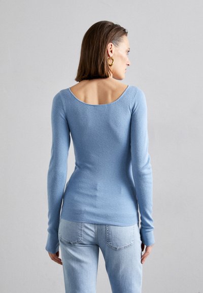 Vaaleansininen muotoiltu pitkähihainen paita pehmeästä neulosmateriaalista, jossa on avara selkäosa ja ribbikuvio, yhdistettynä vaaleansinisiin farkkuihin.