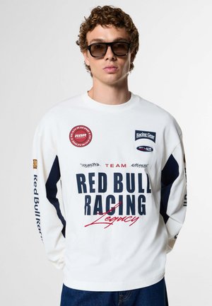 Junger Mann mit lockigem Haar, der schwarze Sonnenbrillen und ein weißes Langarmshirt mit Red Bull Racing-Logos und dunkelblauen Akzenten trägt.