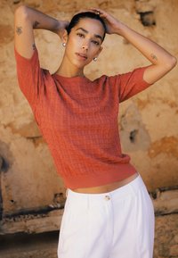 Femme aux cheveux tirés en arrière, portant un haut texturé à manches courtes couleur rouille et un pantalon blanc, posant avec les mains derrière la tête contre un mur usé.