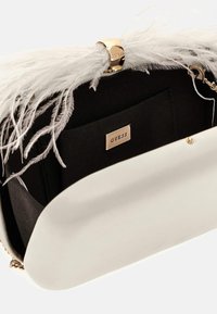 Balto odos delninė su auksiniu užsegimu ir dekoratyvine balta plunksna, juodu interjermu ir ženklo etikete.