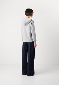 Personne avec les cheveux courts portant un sweat à capuche gris clair et un jean large bleu foncé, se tenant devant un fond blanc uni, de profil.