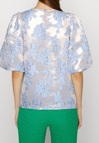 Blusa de color azul claro con estampado floral y mangas abullonadas, hecha de tela brillante. Cierre en la parte trasera con un ojal. Llevada con pantalones verdes con textura.