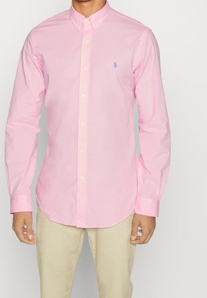 Mężczyzna ubrany w dopasowaną, jasnoróżową koszulę zapinaną na guziki oraz beżowe spodnie, stojący z rękami swobodnie opuszczonymi wzdłuż boków na jednolitym tle.