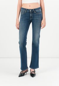 Sieviete, tērpusies vidus augstuma zilās bootcut džinsos un melnās norādītā pirksta slēgšanas kurpēs, stāv uz baltā fona.