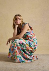 Sieviete čurā, atpūšot galvu uz vienas rokas, valkājot bezroku kleitu ar volāniem, uz kuras ir spilgtas daudzkrāsainas ziedu raksti uz baltas pamatnes.