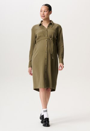 Olivgrünes Hemdkleid mit langen Ärmeln, strukturiertem Stoff und gebundenem Taillengürtel. Das Model trägt schwarze Slipper und weiße, gestreifte Socken.