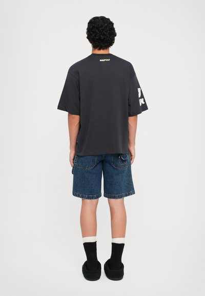 Čierne tričko s krátkym rukávom a logom "NAPOLI" na chrbte, skombinované s modrými džínsovými šortkami. Model má na sebe čierne topánky apono biele pruhované ponožky.