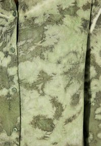 Zelená košile s knoflíkovým zapínáním vyrobená z lehkého materiálu s tie-dye vzorem, s tmavšími zelenými akcenty a náprsní kapsou. Knoflíky jsou kulaté a neprůhledné.