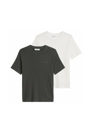 Hombre con camiseta blanca lisa de manga corta, vaqueros azul claro, collar de cadena de plata y pulsera, con una mano en el bolsillo.