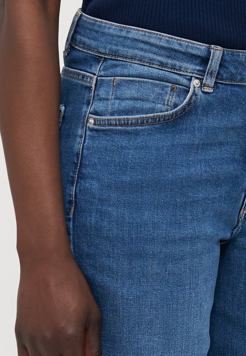 Jean en denim bleu moyen, présentant un design classique à cinq poches, une coupe slim et un léger délavage. Détails de couture le long des coutures.