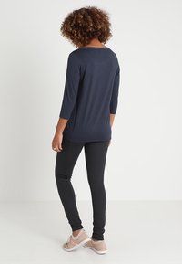 Frau mit lockigem Haar, die ein marineblaues Langarmoberteil, schwarze enganliegende Hosen und beigefarbene Turnschuhe trägt, steht vor einem weißen Hintergrund.