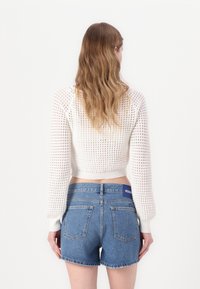 Hvit strikket croppgenser med lange ermer og mønstret tekstur, kombinert med blå denimshorts med en logo-patch på baksiden.