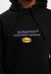 Felpa nera con cappuccio e testo viola, con la scritta "JACK&JONES NINETEEN NINETY" e un logo giallo "Originals". Texture morbida, tasca frontale.