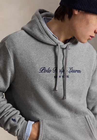 Une personne portant un sweat à capuche gris Polo Ralph Lauren avec une inscription bleu marine et un bonnet bleu marine, une main dans la poche avant.