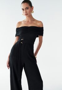 Black off-the-shoulder jumpsuit with textured fabric, fitted bodice, wide legs, and a gold-tone belt buckle accent.