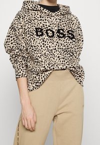 Beiger Hoodie mit schwarzem Leopardenmuster und in Schwarz gesticktem "BOSS". Mit langen Ärmeln und Kapuze mit Kordelzug. Kombiniert mit beigen Hosen.