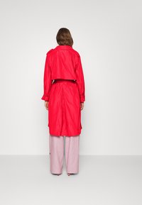 Czerony wodoodporny płaszcz trench z paskiem w talii, długimi rękawami i skróconym krojem, w połączeniu z jasnoróżowymi spodniami typu wide-leg.